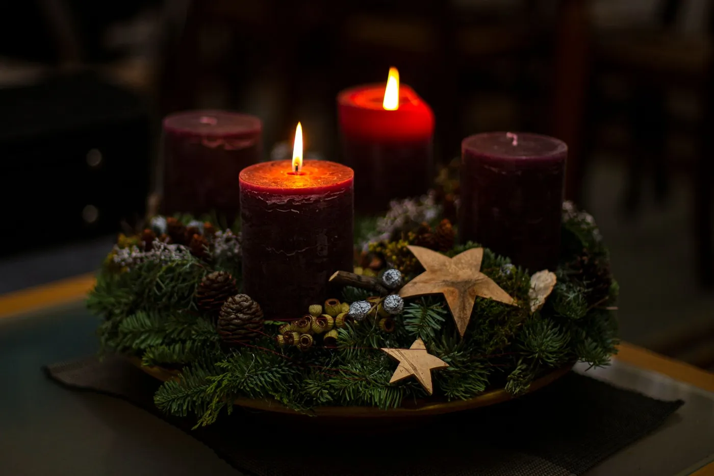 An Advent wreath on a table with two of its four candles lit, resting on evergreen branches with pine cones and wooden stars
