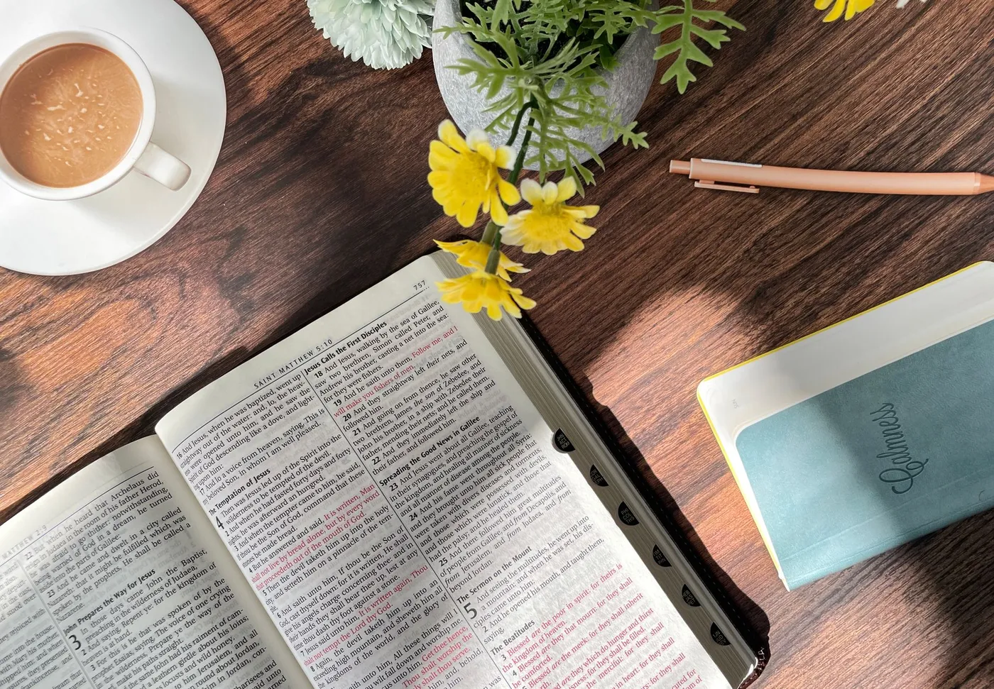 An open Bible at Matthew 4 and 5 on a wooden table in morning sunlight, beside a cup of coffee, yellow flowers, a pen and a small notebook