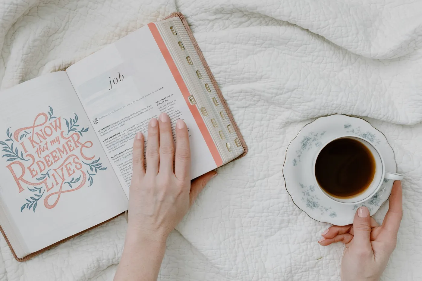 An open Bible resting on a white quilt beside a cup of coffee, with a hand holding the page open at the book of Job.