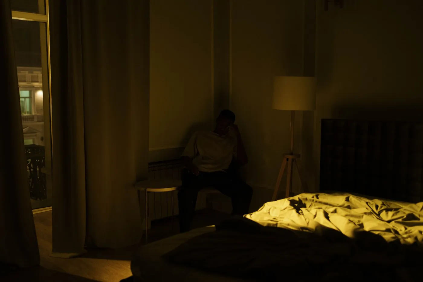 A person sitting alone on a bench in a dark bedroom at night, lit only by a floor lamp, with an unmade bed in the foreground