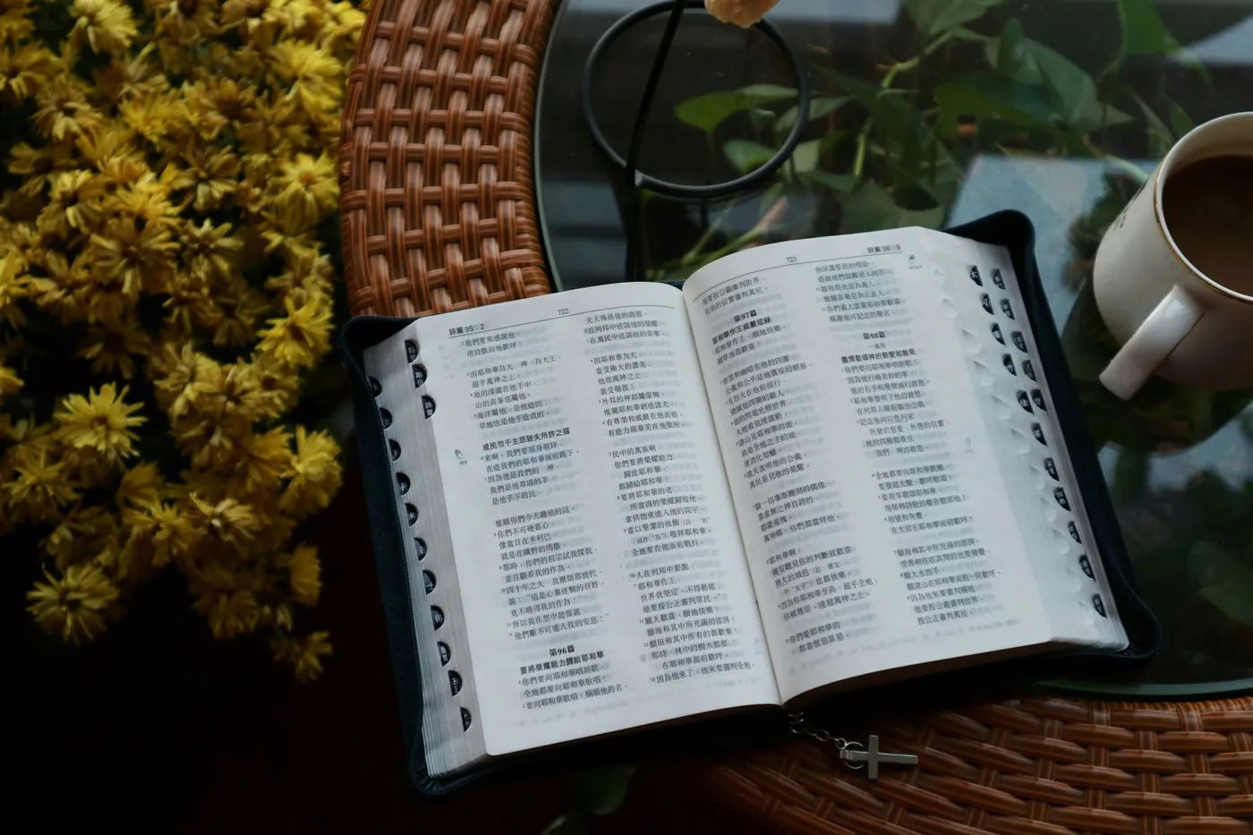 A Bible lying open to the Psalms on a table beside a mug and a spray of yellow flowers, its ribbon and small cross bookmark resting across the page