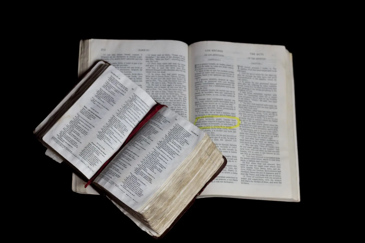 Two open Bibles on a black background, the larger one turned to the page where the Gospel of John ends and the Acts of the Apostles begins