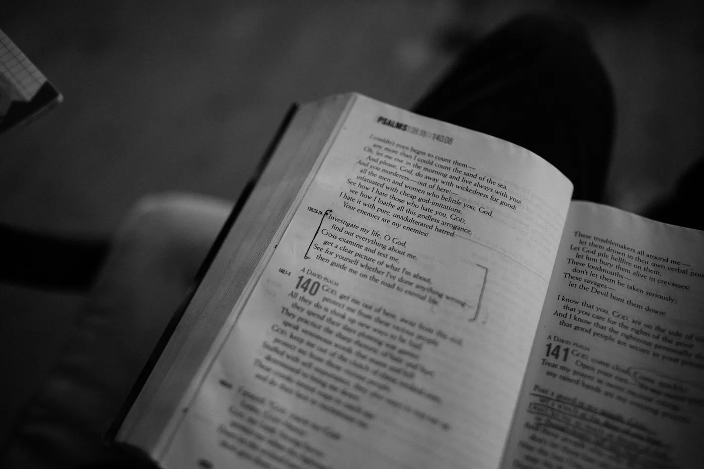 A black and white photo of an open Bible held on a reader's lap, turned to the end of Psalm 139 and the start of Psalms 140 and 141, with Psalm 139:23-24 bracketed in pen