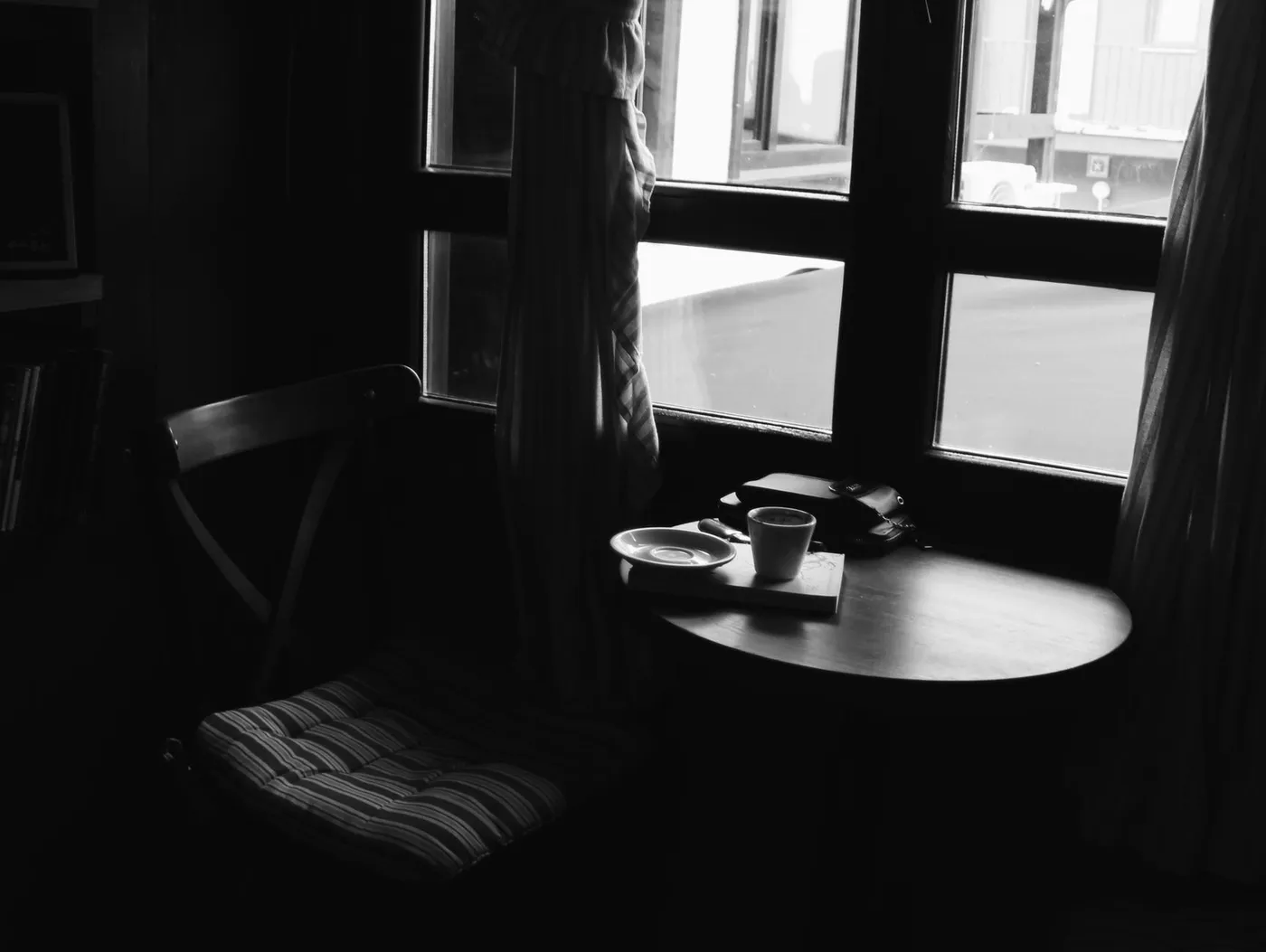 An empty chair at a small round table by a window, with a coffee cup, saucer and a closed notebook on it, in black and white