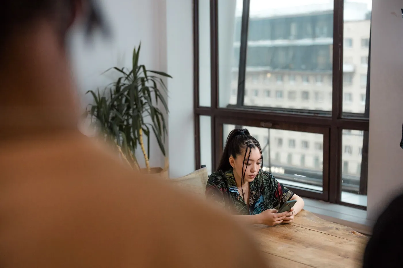 A woman looks at her phone at a wooden dining table while another person sits across from her, out of focus in the foreground