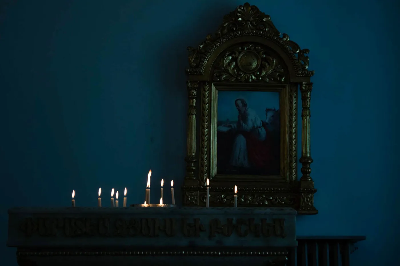 Votive candles burning on a stone ledge below an icon in a gilded frame, in a dim church interior