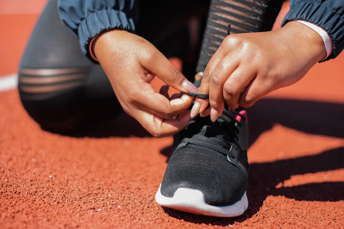 Two hands tying the lace of a black running shoe on an orange athletics track in bright sunlight