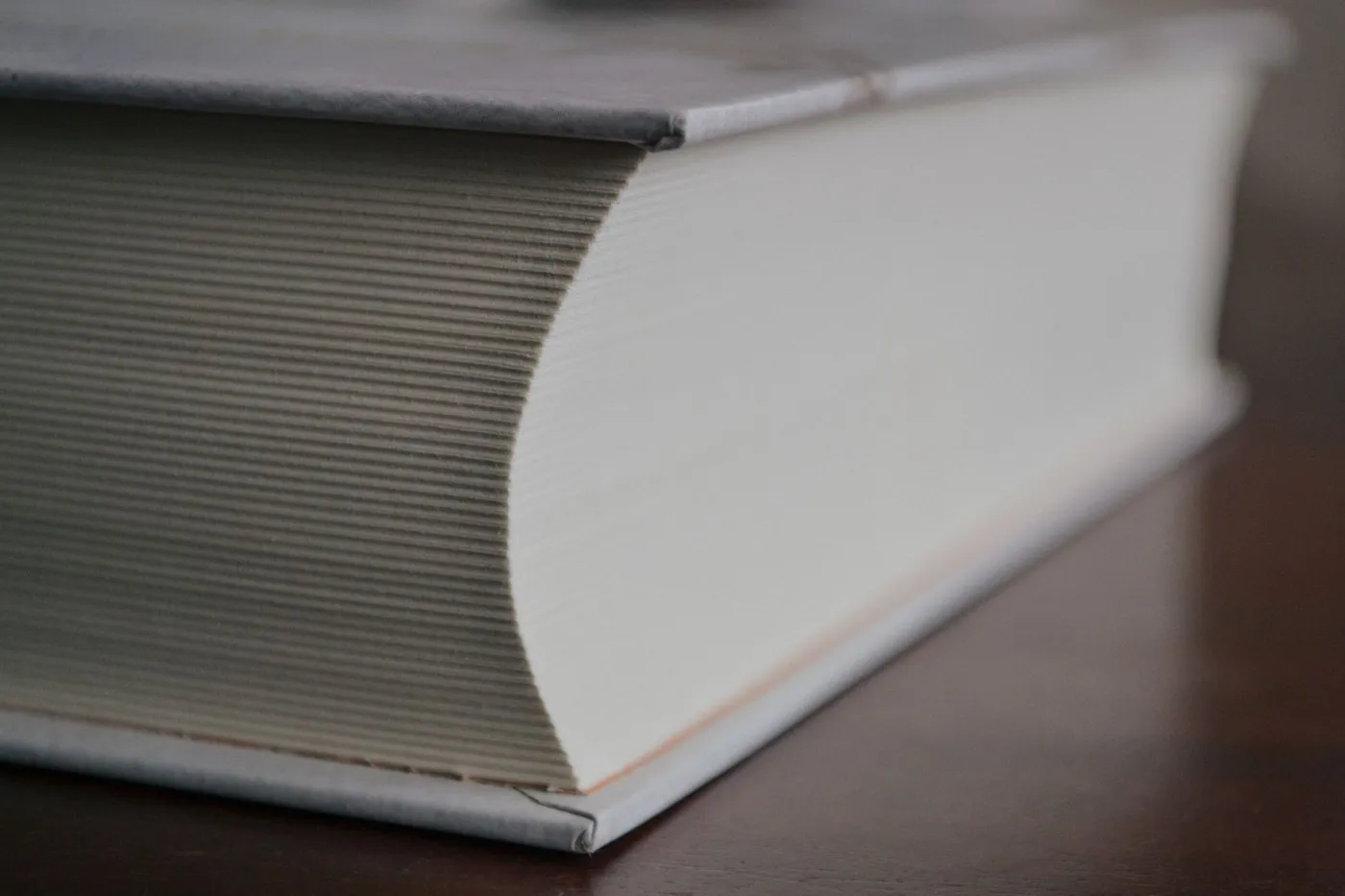 The corner of a thick closed hardback book resting on a dark wooden table, its many page edges visible in close focus