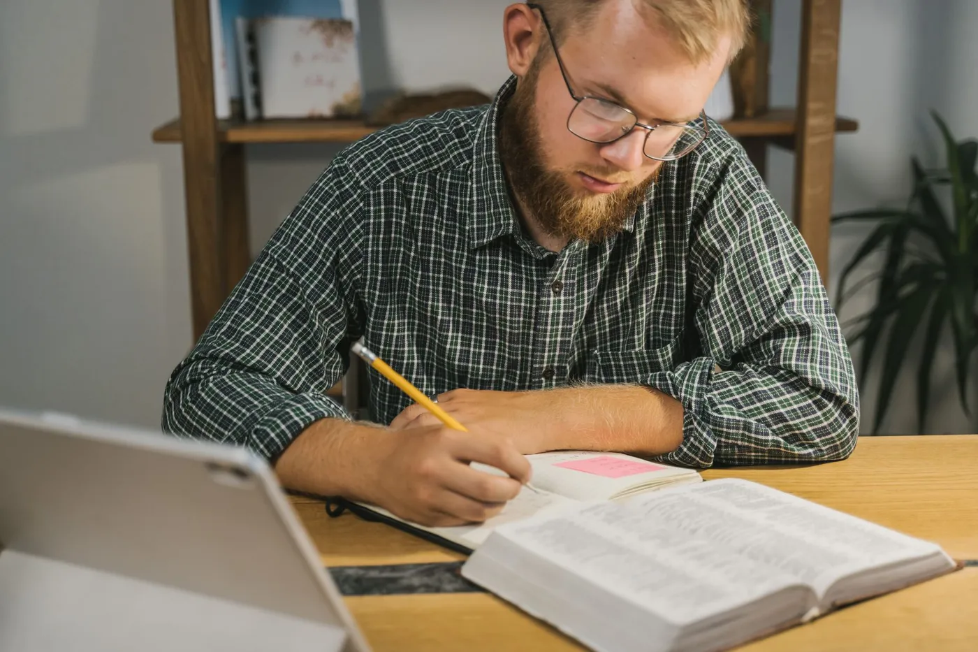 A man in a checked shirt reading an open Bible at a wooden desk and writing notes in a notebook with a pencil, a tablet pushed to the edge of the table
