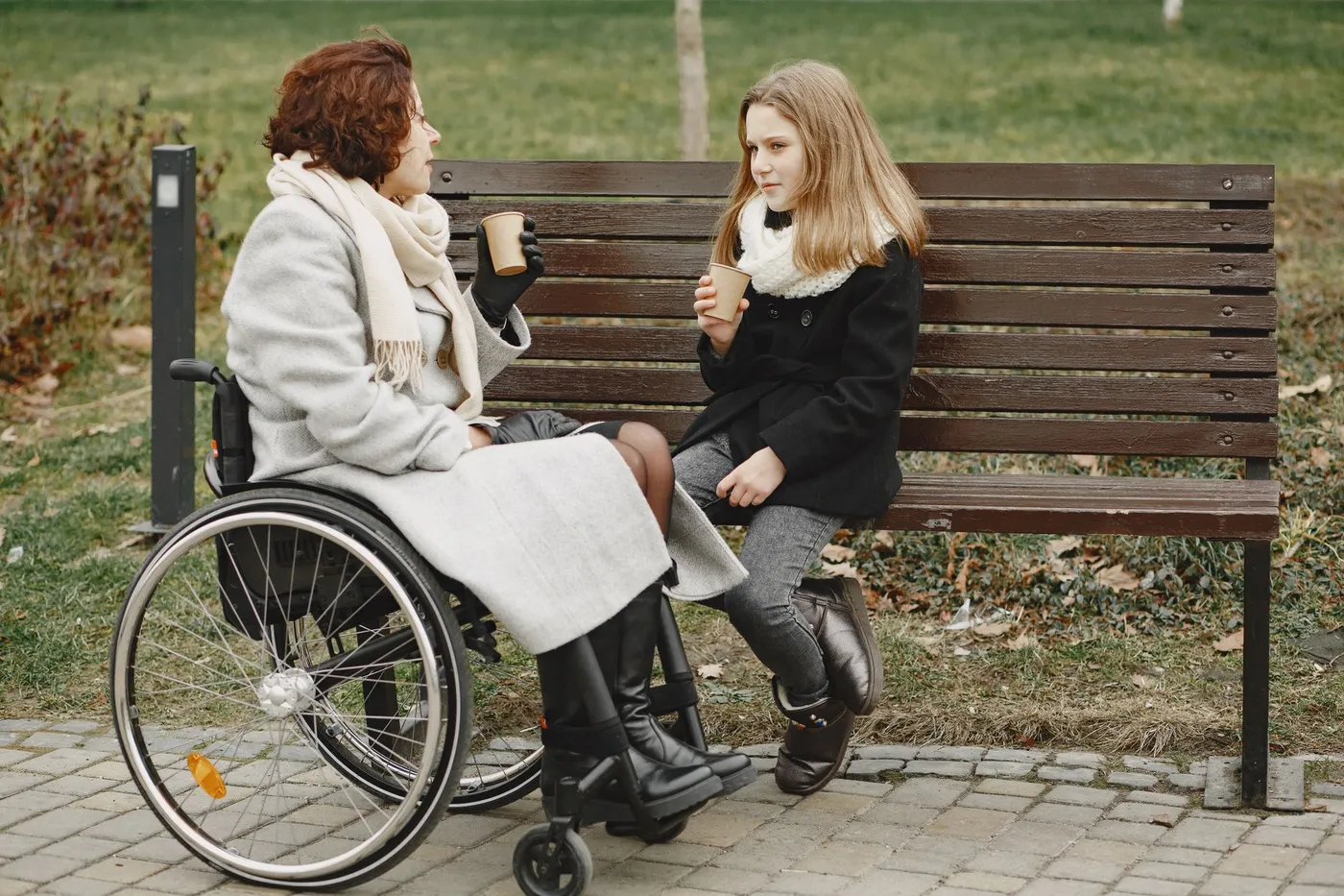 A woman in a wheelchair and a girl on a park bench, each holding a paper cup, in a serious conversation
