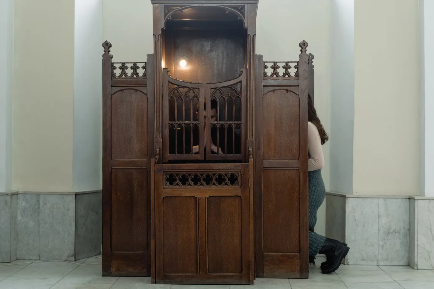 A carved wooden confessional in a church, with a priest seated behind a latticed grille and a woman kneeling at the screen on the right side