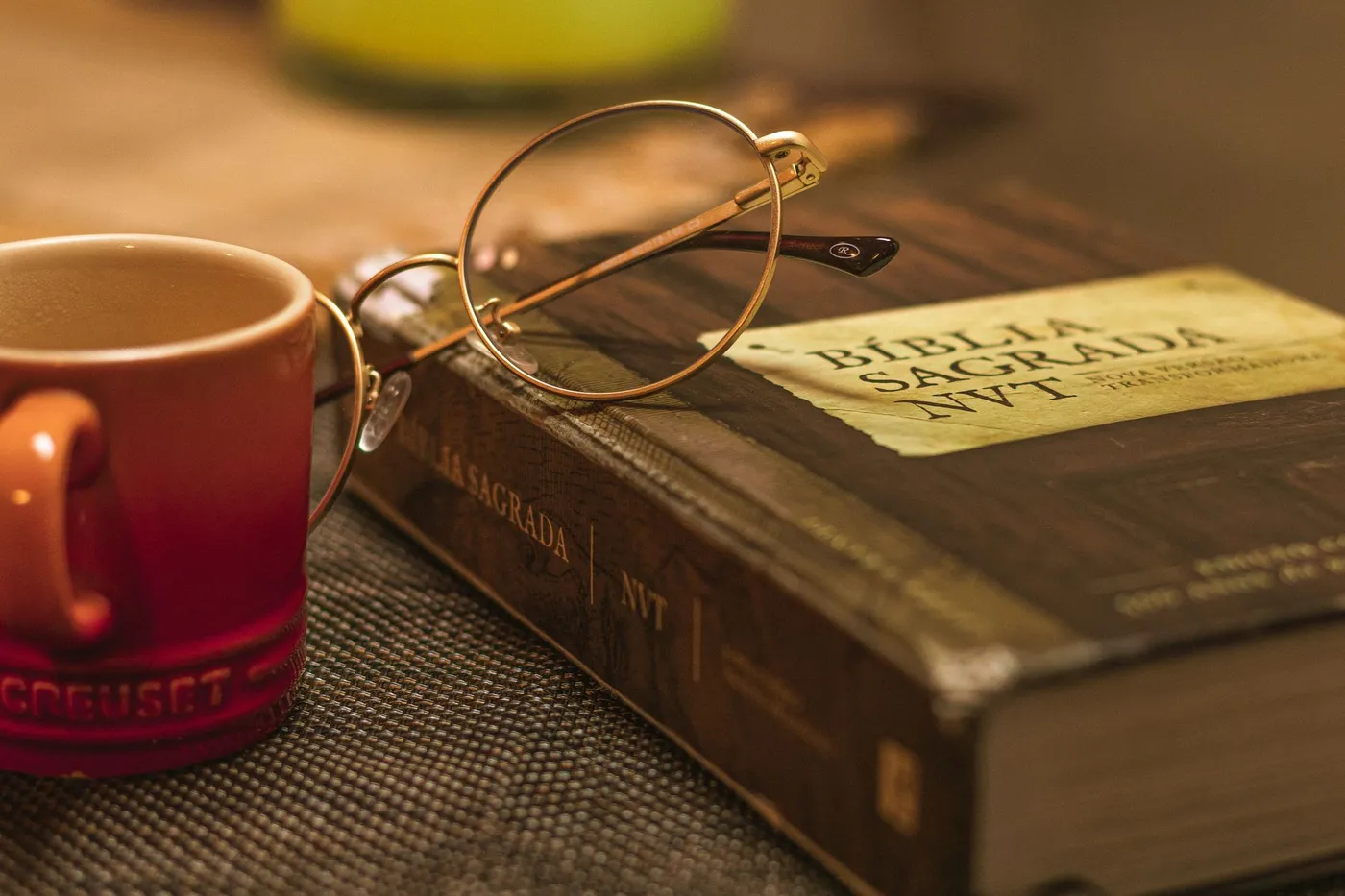 A closed Portuguese-language Bible with a pair of round wire glasses resting on it, beside a red coffee mug on a woven placemat