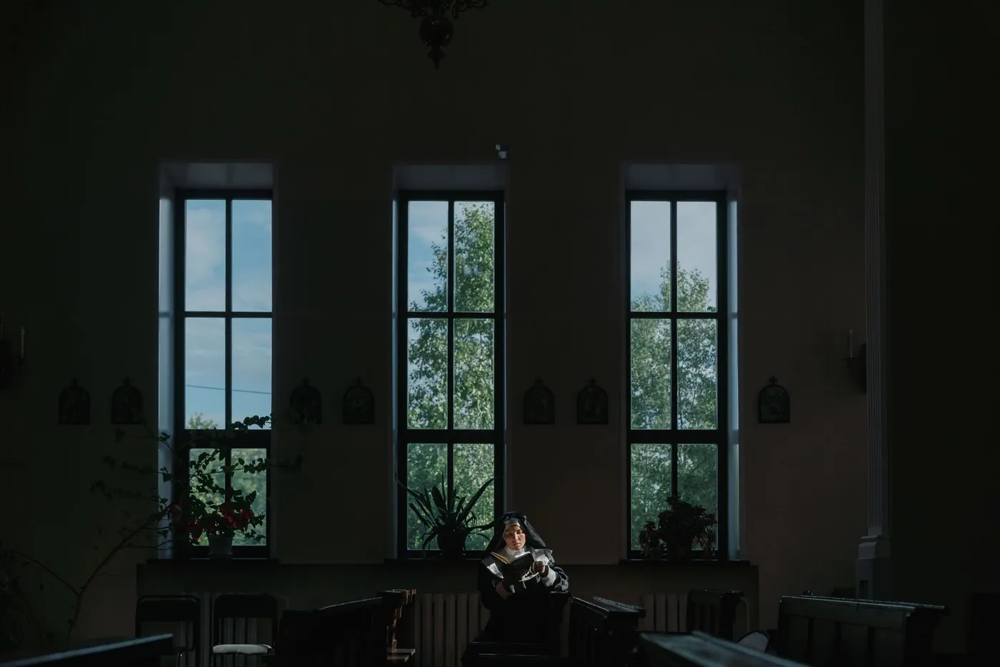 A religious sister in a black habit reading a book alone in a dim church, lit by three tall windows with green trees outside