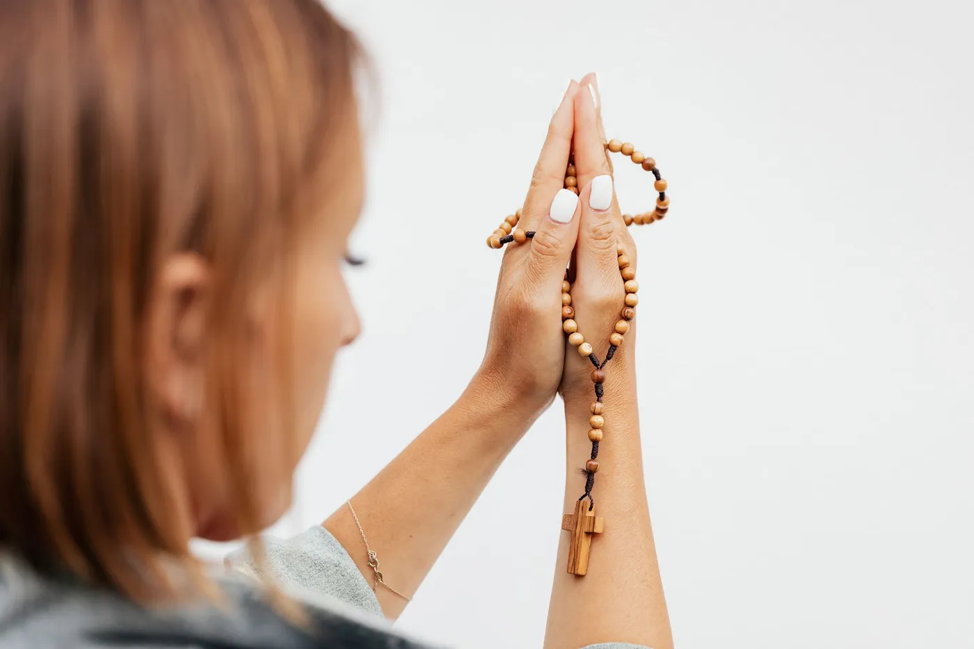 A woman seen from behind holding a wooden rosary between her joined hands, the small wooden cross hanging below her wrists
