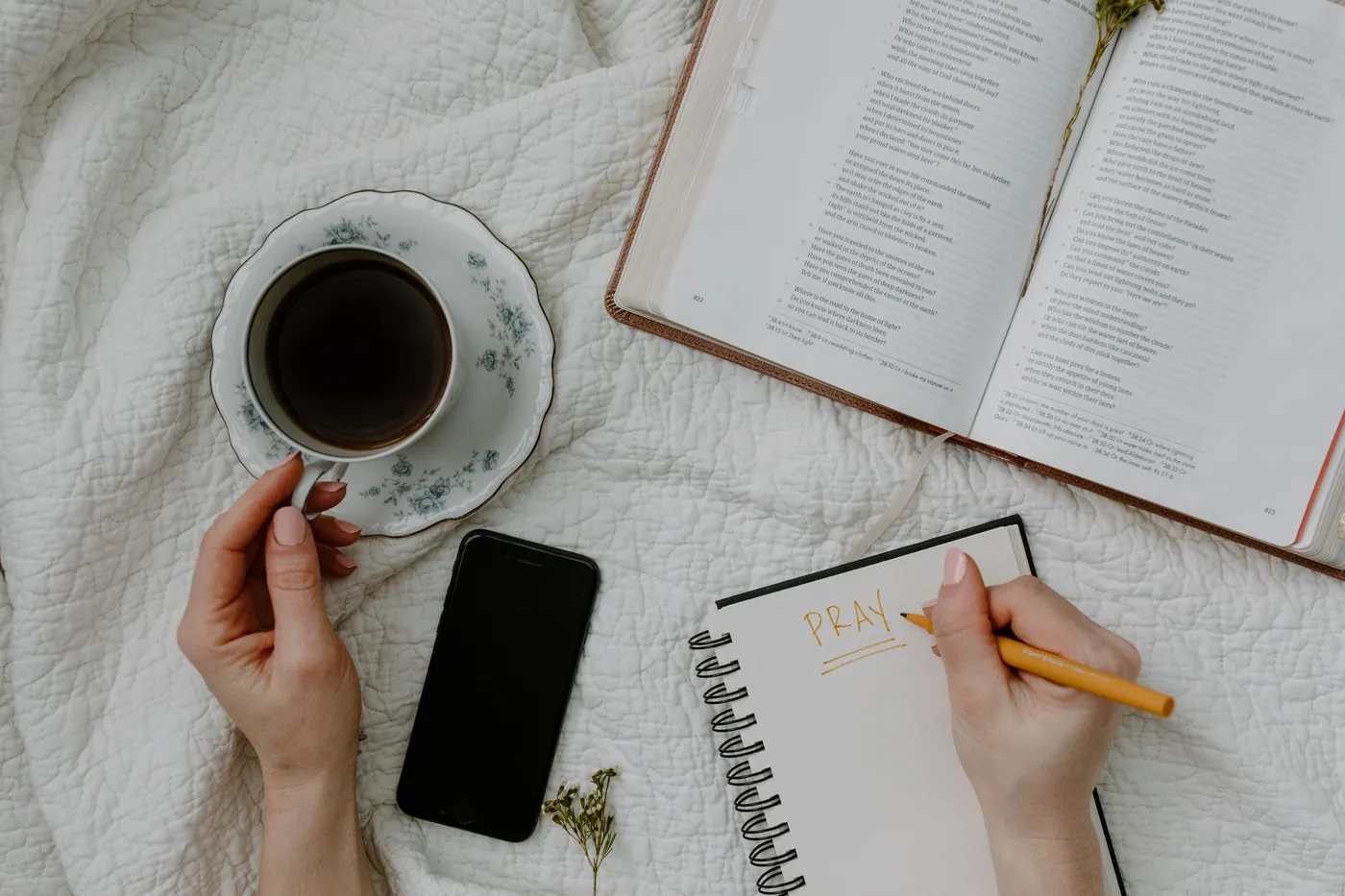An open Bible, a spiral notebook with the word PRAY written on it, a cup of coffee and a phone lying face down on a white quilt