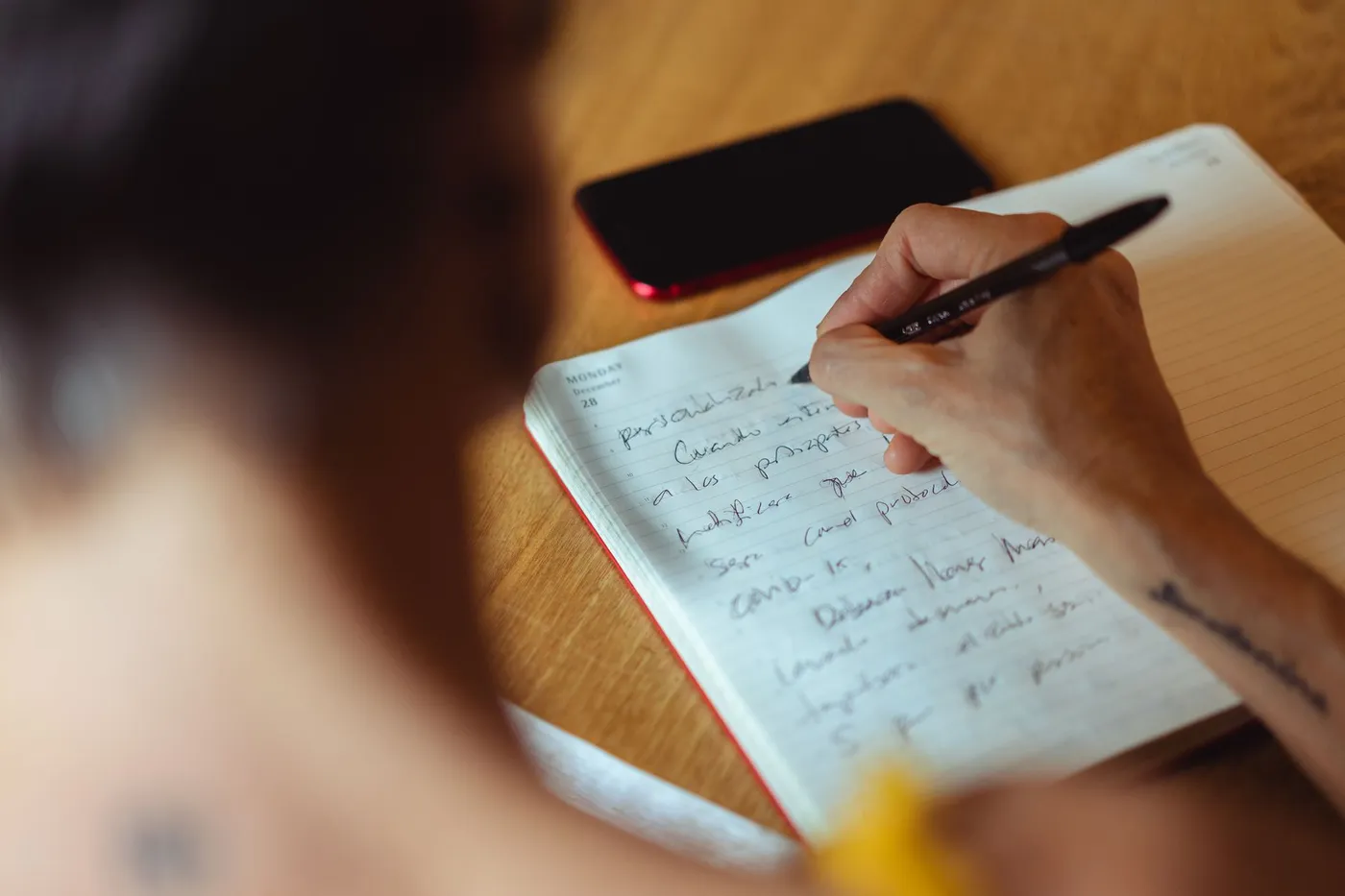 A person writing by hand in a lined notebook at a wooden desk, with a smartphone lying beside the page