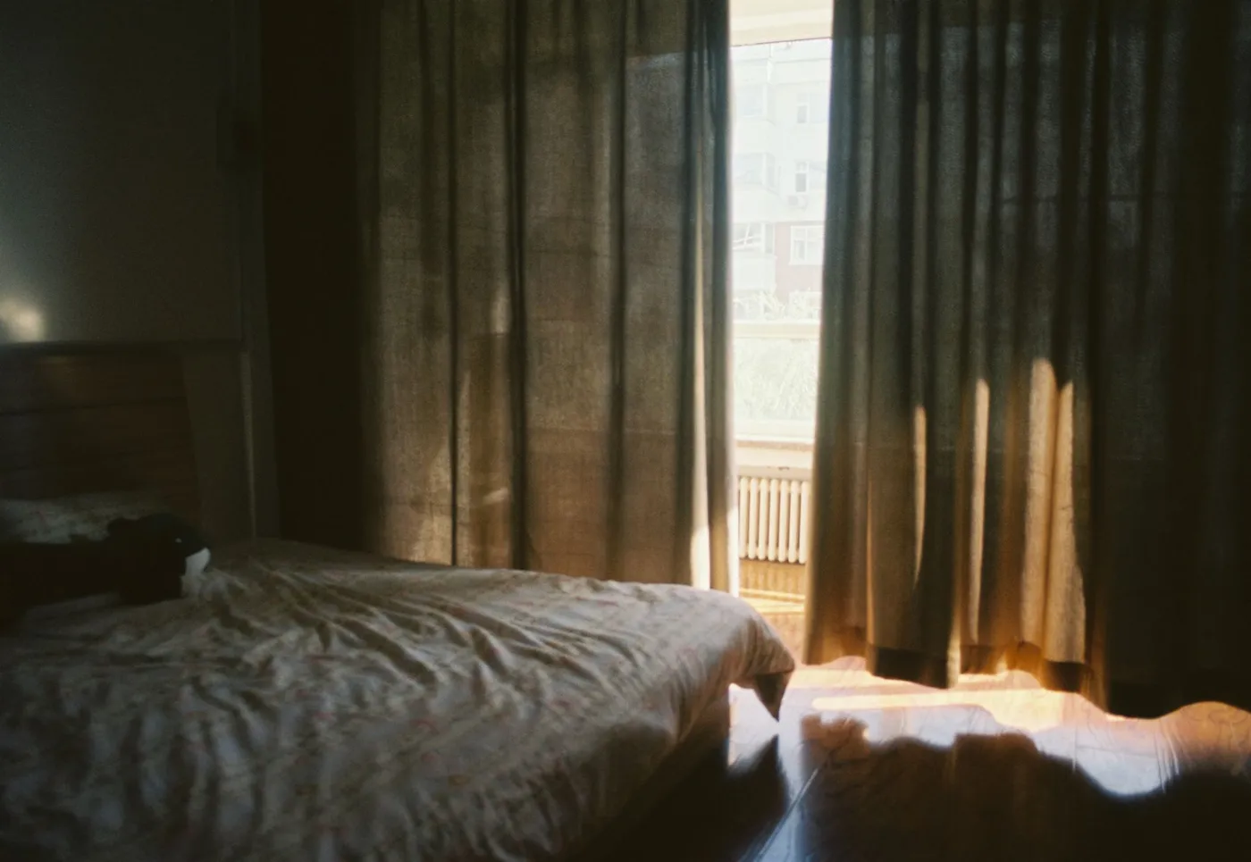 A bedroom in early morning light, curtains half drawn over an unmade bed