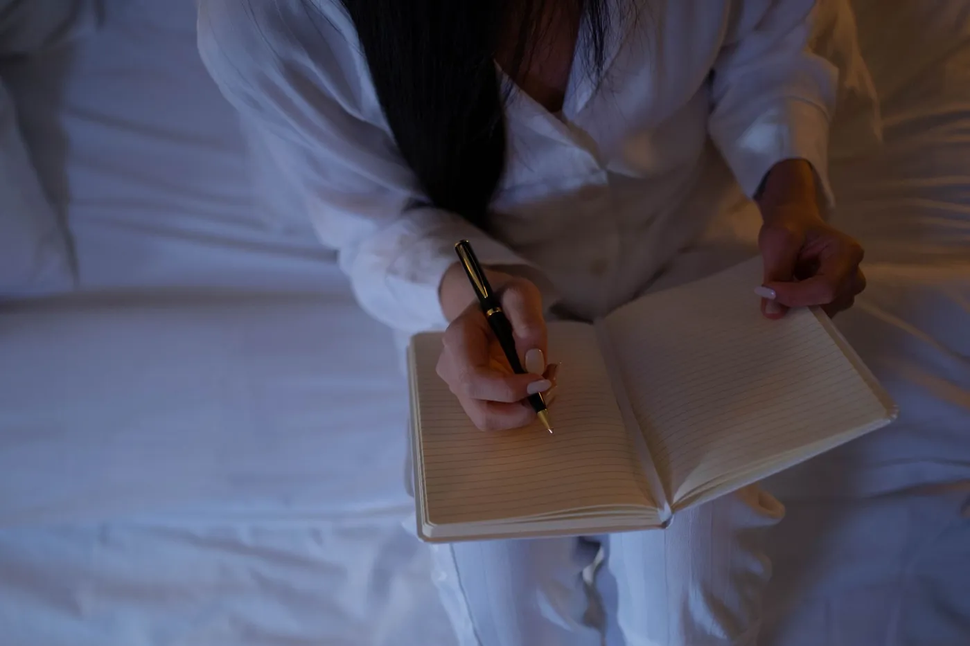 A person in white pajamas sitting on a bed in low evening light, writing with a black pen in an open lined notebook
