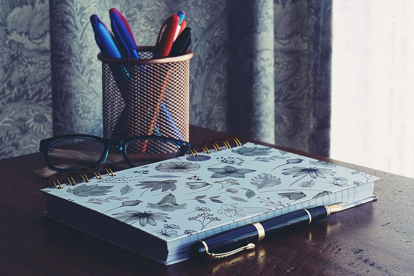 An open notebook with blank ruled pages lying on a desk beside several pens.