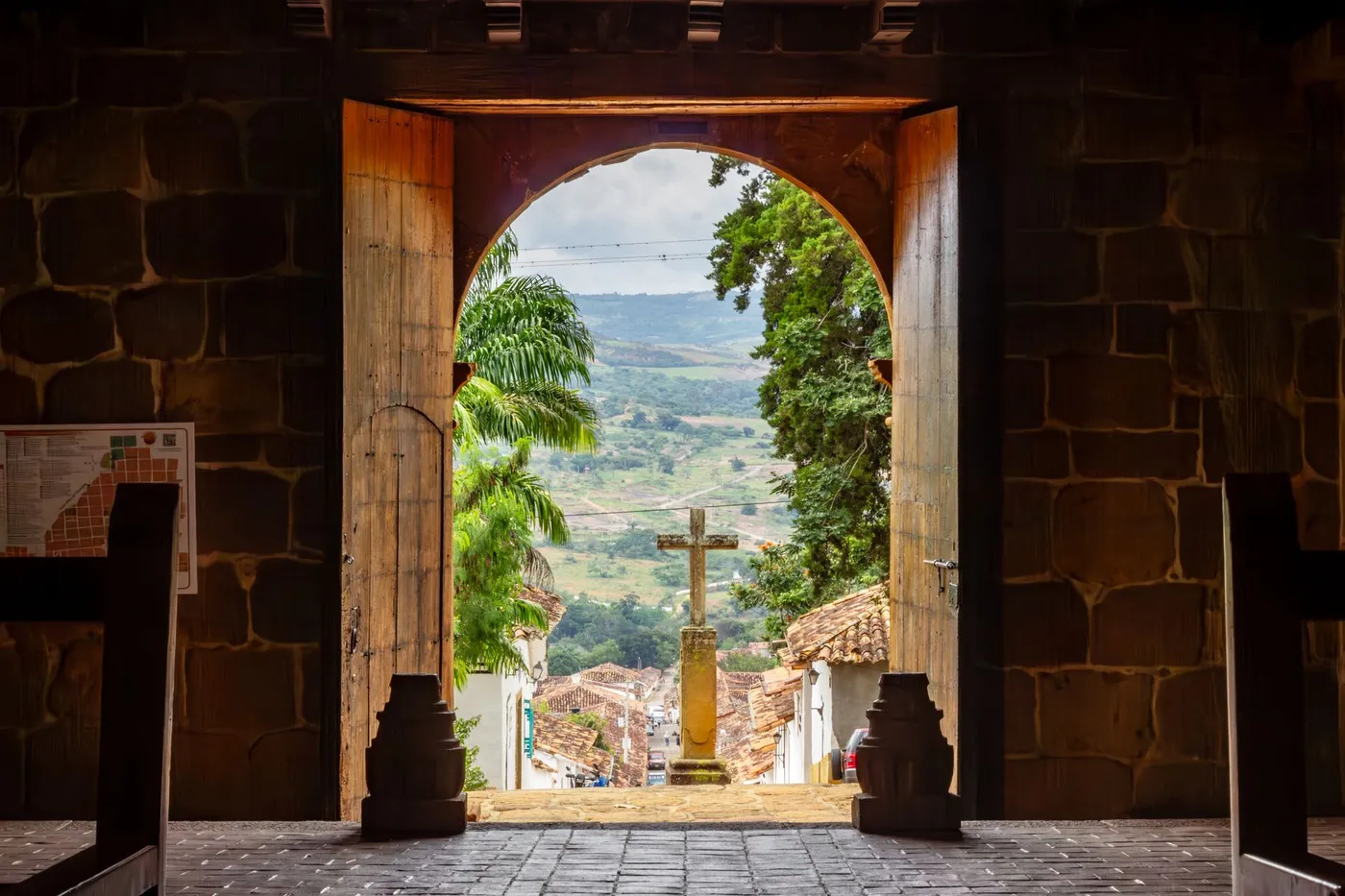 The open wooden doors of a stone church seen from inside, looking out onto a stone cross, tiled rooftops and green hills beyond