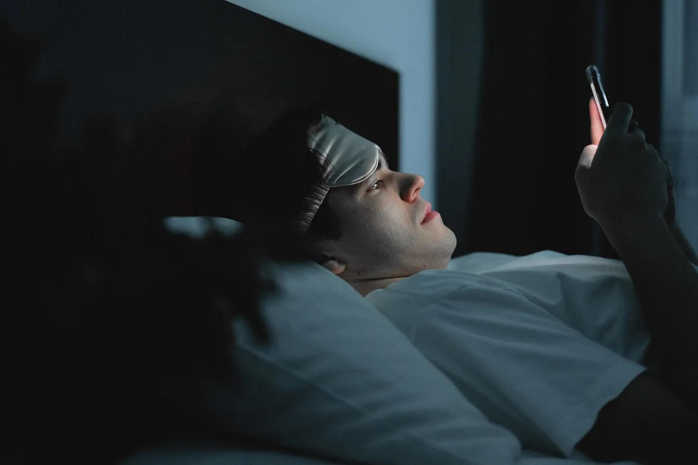 A man lying on his side in bed at night, his face lit by the screen of the phone he is holding.