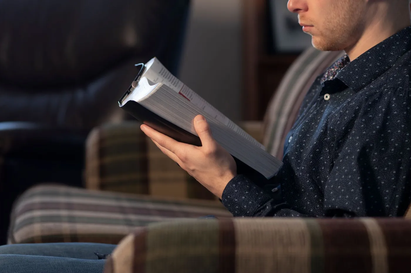 A man sitting in an armchair holding open a thick hardback book with a ribbon marker