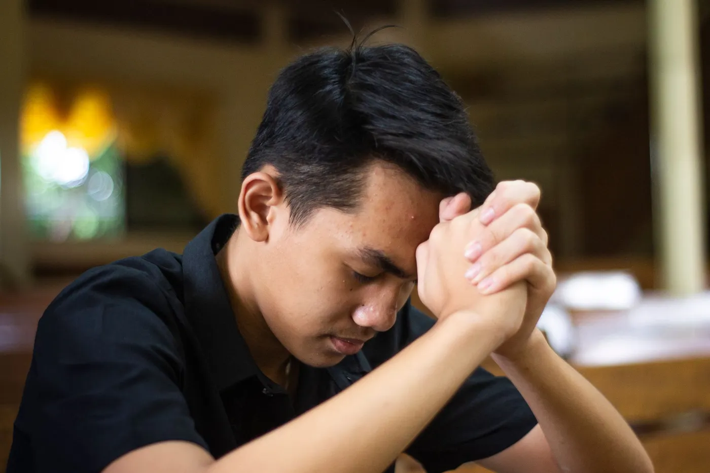 A young man with his eyes closed and his hands clasped against his forehead, praying inside a church