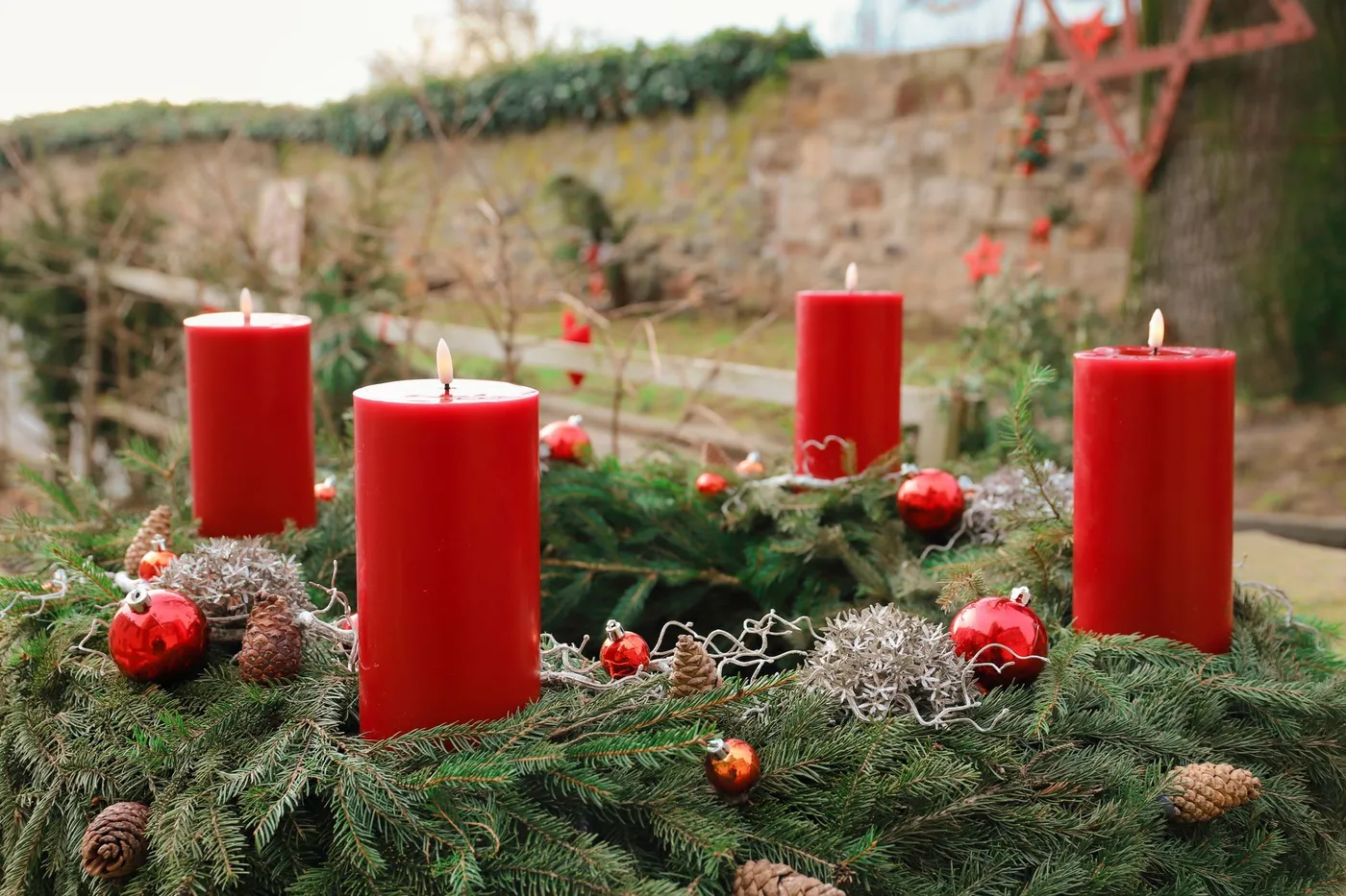 An Advent wreath of evergreen branches holding four red candles, set outdoors among pine cones and red ornaments.