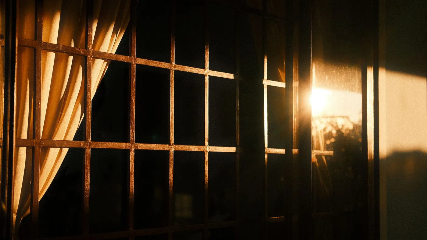 Warm early sunlight coming through a barred window and a pale curtain into a dark room