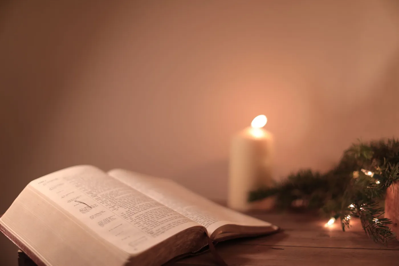 An open Bible on a wooden table beside a lit candle and a sprig of pine with Christmas lights