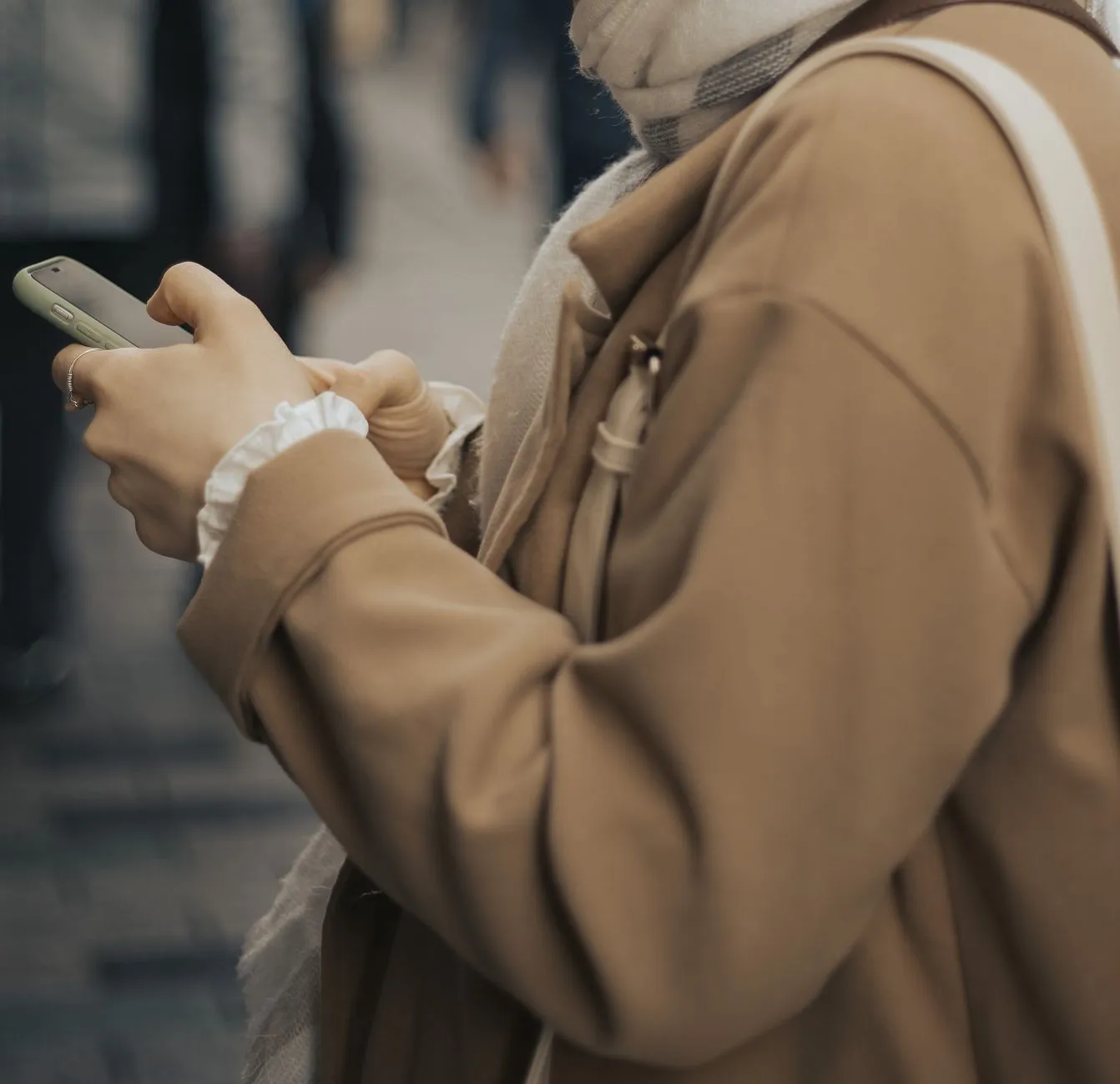 A person in a camel coat and scarf standing on a busy street, looking down at a smartphone held in both hands