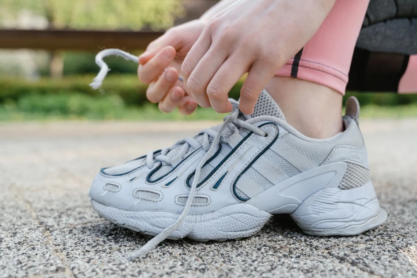 A person crouched on a paved path tying the lace of a grey running shoe, with a green park blurred behind